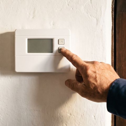 A hand presses a button on a plain wall thermostat in a plaster hallway, beside a coat on a hook